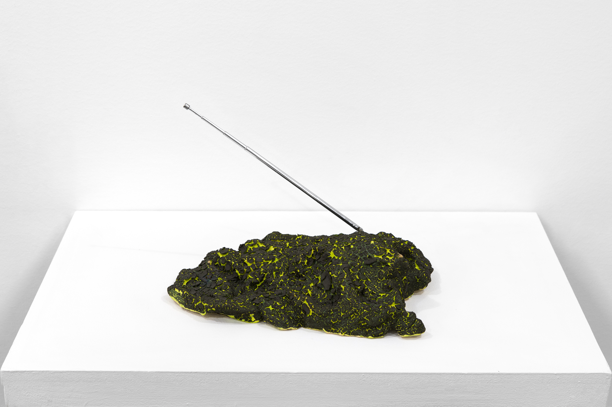 A green object with a cracked, black surface sits on a white base. A metal antenna extends out on a diagonal.