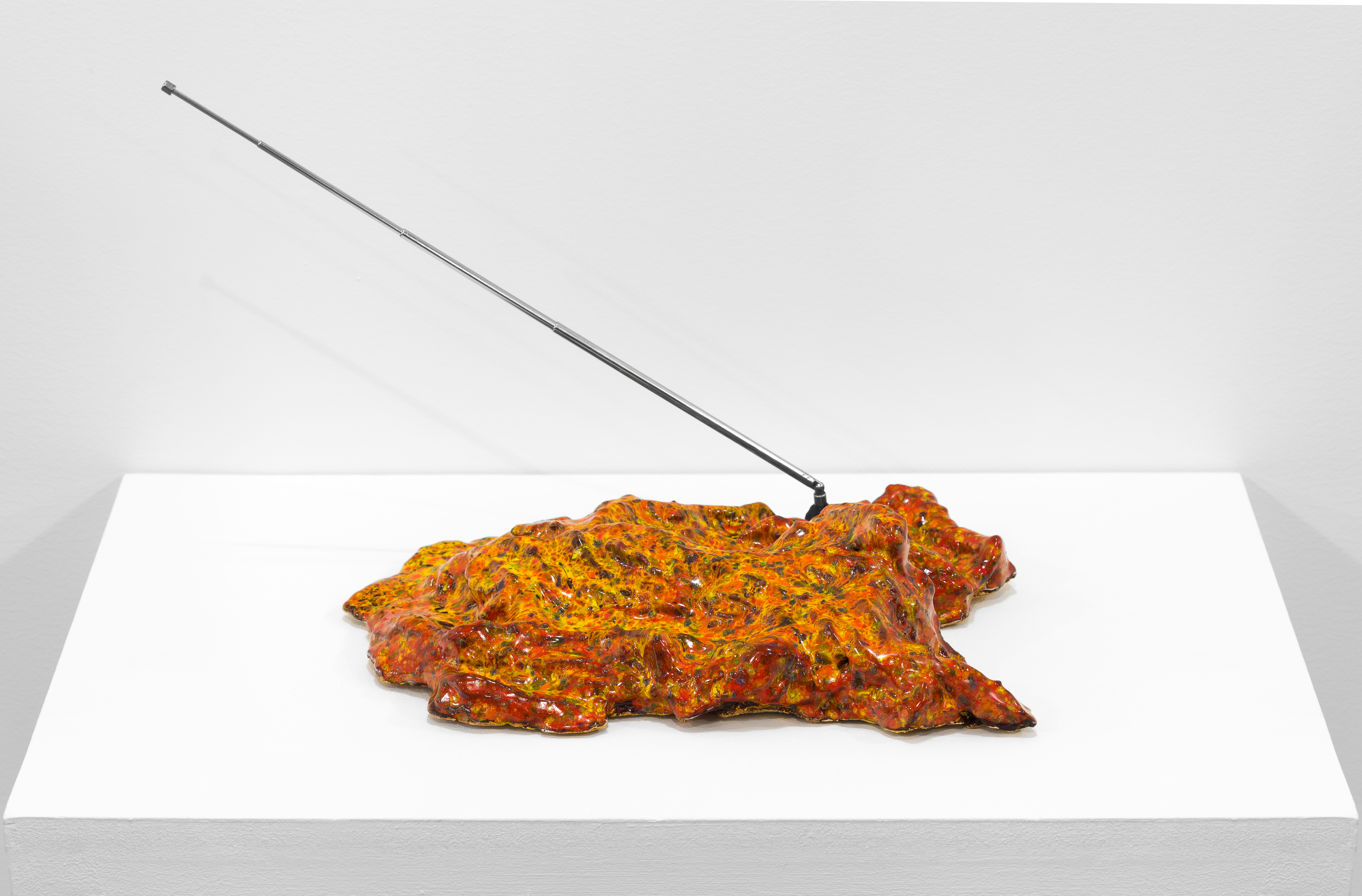 A multicolored object sits on a white base. A metal antenna extends out on a diagonal.