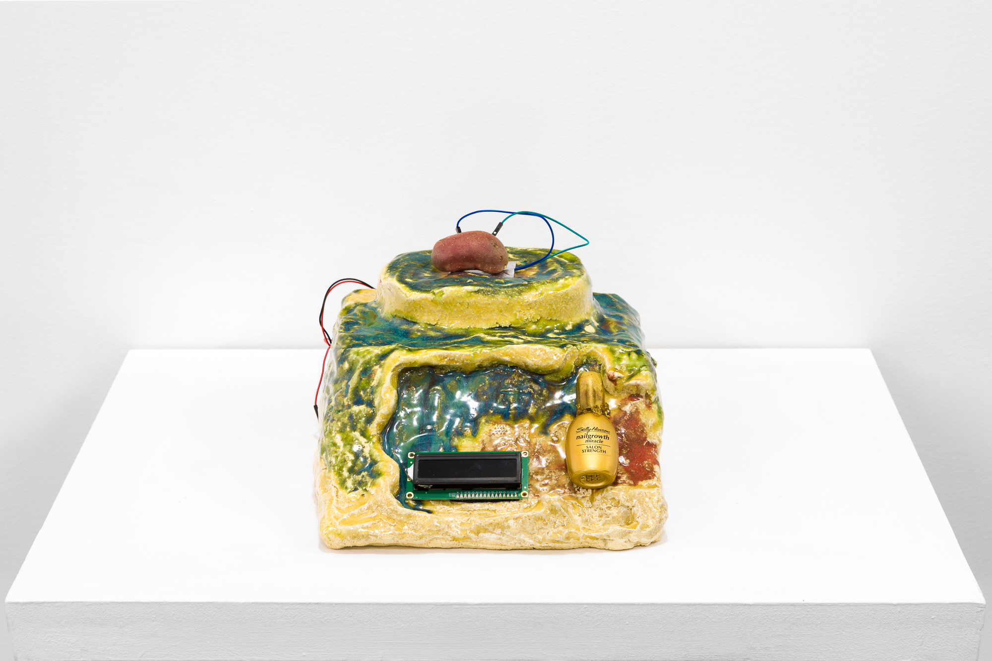 A yellow and blue object sits on a white base. Embedded in the front of the object is black electronic screen and a gold nail polish bottle reading 