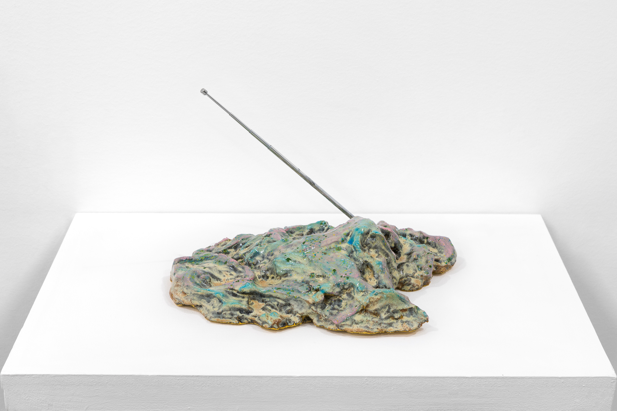 An object with tones of tan, blue, pink, and grey sits on a white base. A metal antenna extends out on a diagonal.