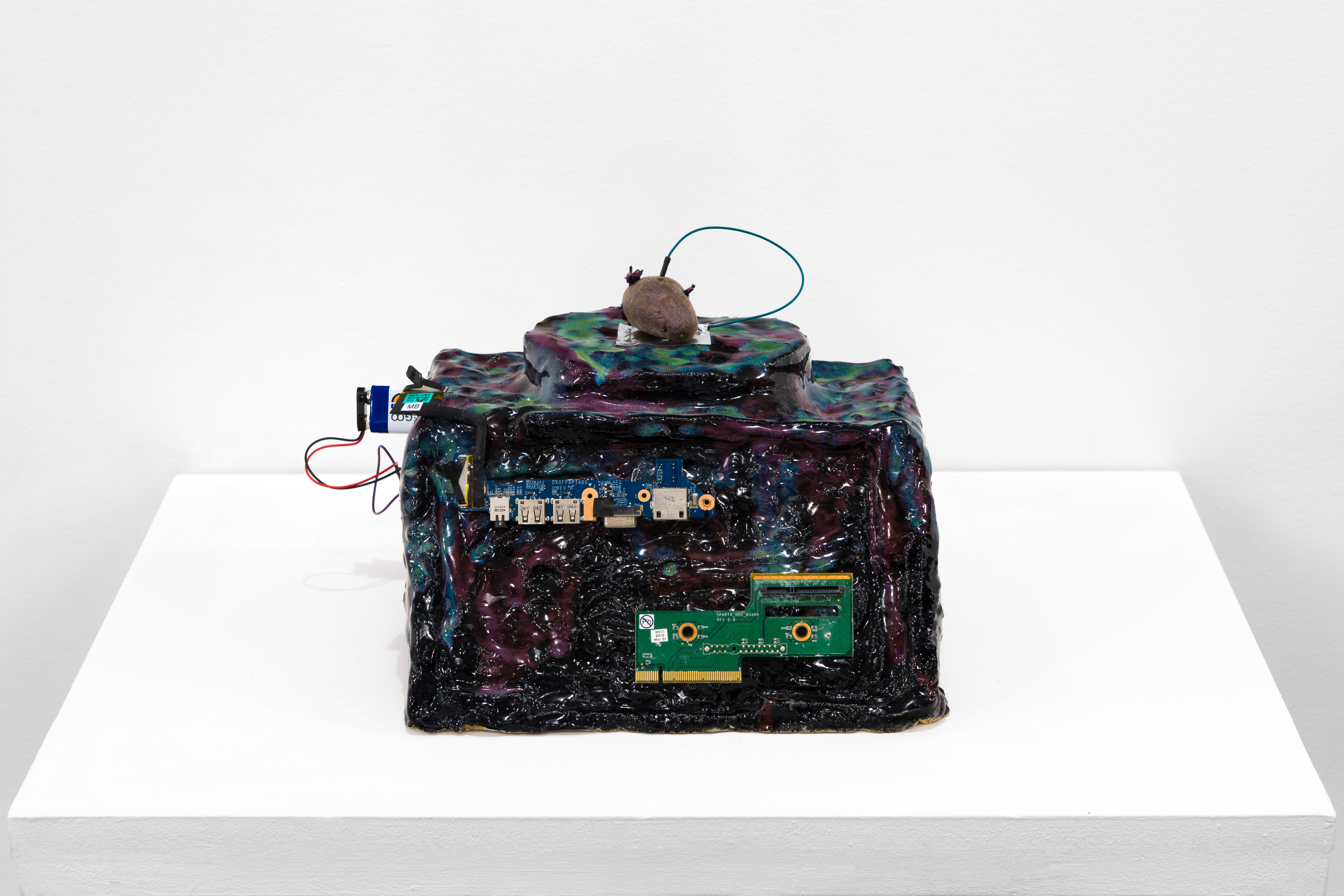 An abstract, multicolored squareish sculpture standing on a white base. A potato sits on top of the sculpture with a wire plugged into the top. A 9v battery is affixed to the side of the sculpture, connected to two comptuer chips affixed to the front.