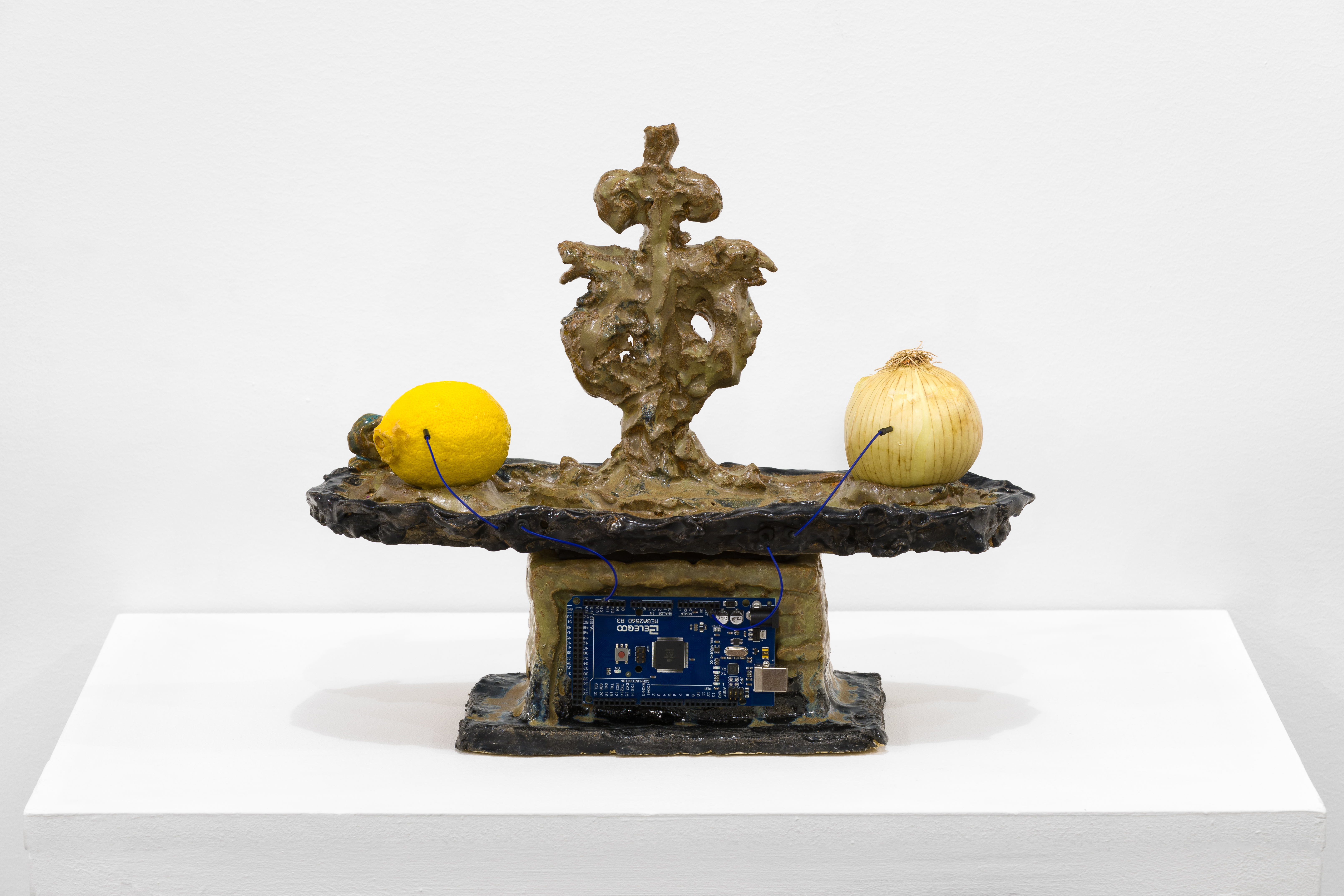 An Elegoo microcontroller is affixed to the side of a brownish sculpture sitting on a white base. Two blue wires are plugged into a lemon and an onion on a platform, each connected to the microcontroller.