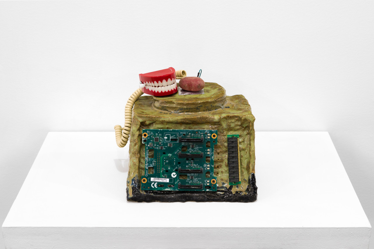 A green object with a processing chip embedded in its front sits on a white base. On top of the object sit a red potato with a wire attached, false teeth, and a telephone cord.