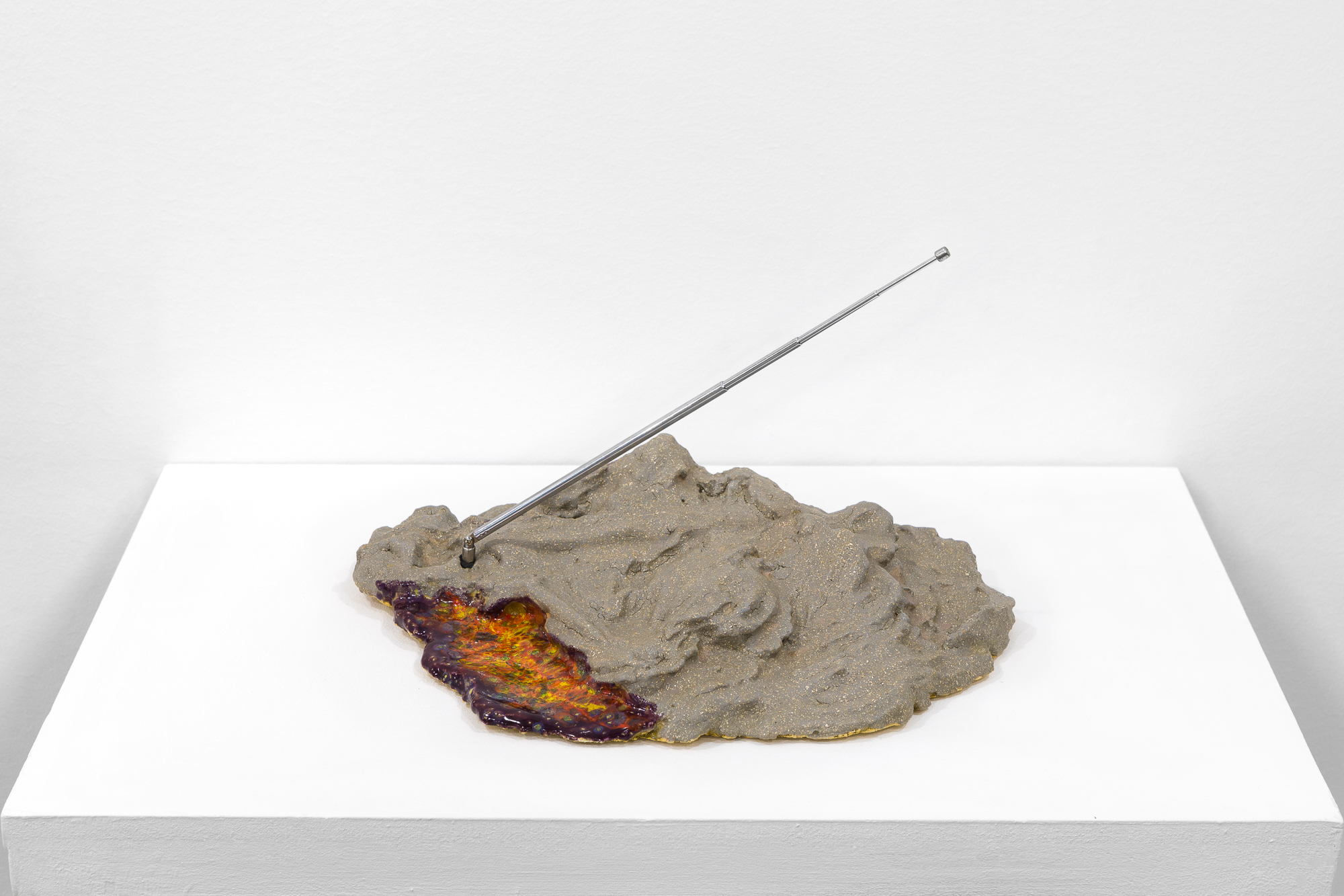 A silver and gold object sits on a white base. A flat, multicolored surface is attached to the left side, and a metal antenna extends out on a diagonal.