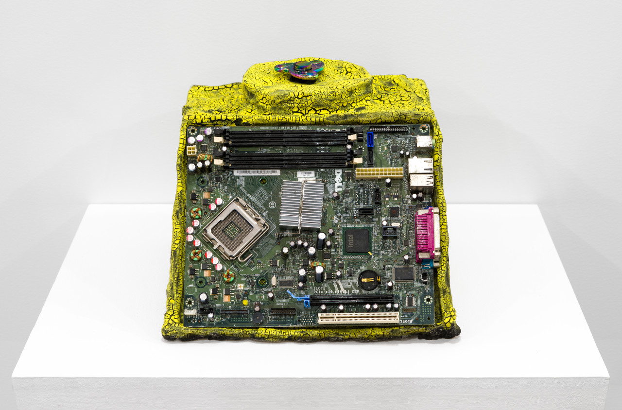 A bright yellow object with a cracked surface is embedded with a computer chipboard on its front, and a multicolored fidget spinner on the top.