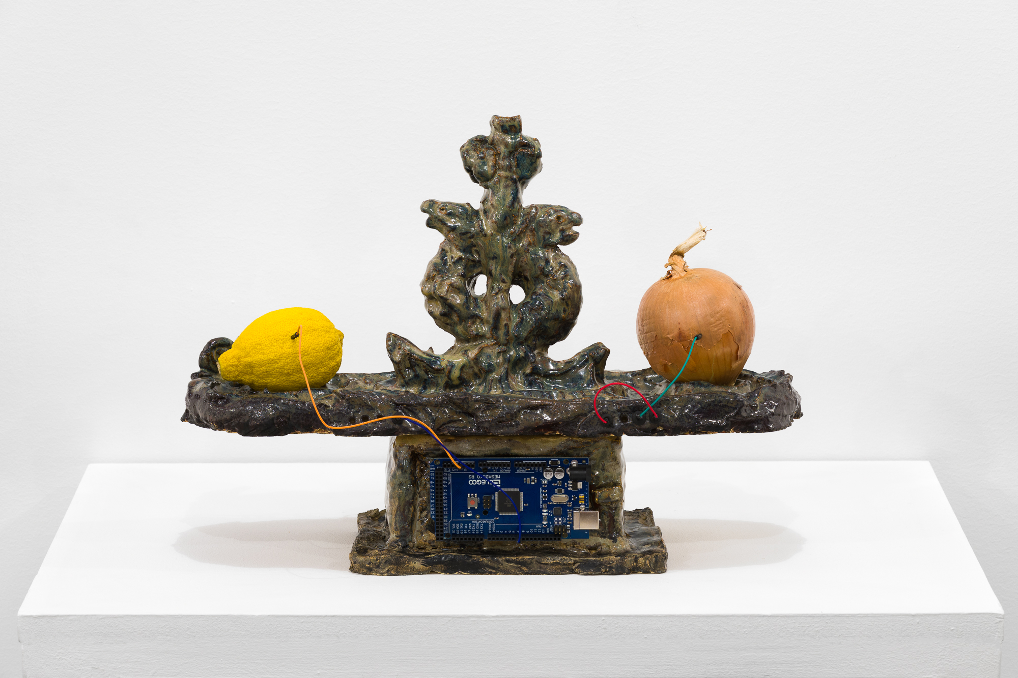 A brown and tan object stands on a white base. On the flat surface of the object, an onion and a lemon sit connected by a rainbow wire. In the base of the object, a processing chip is embedded.