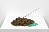 A brown and green object sits on a white base. A blue flat surface is attached to right side of the object, and a metal antenna extends on a diagonal.
