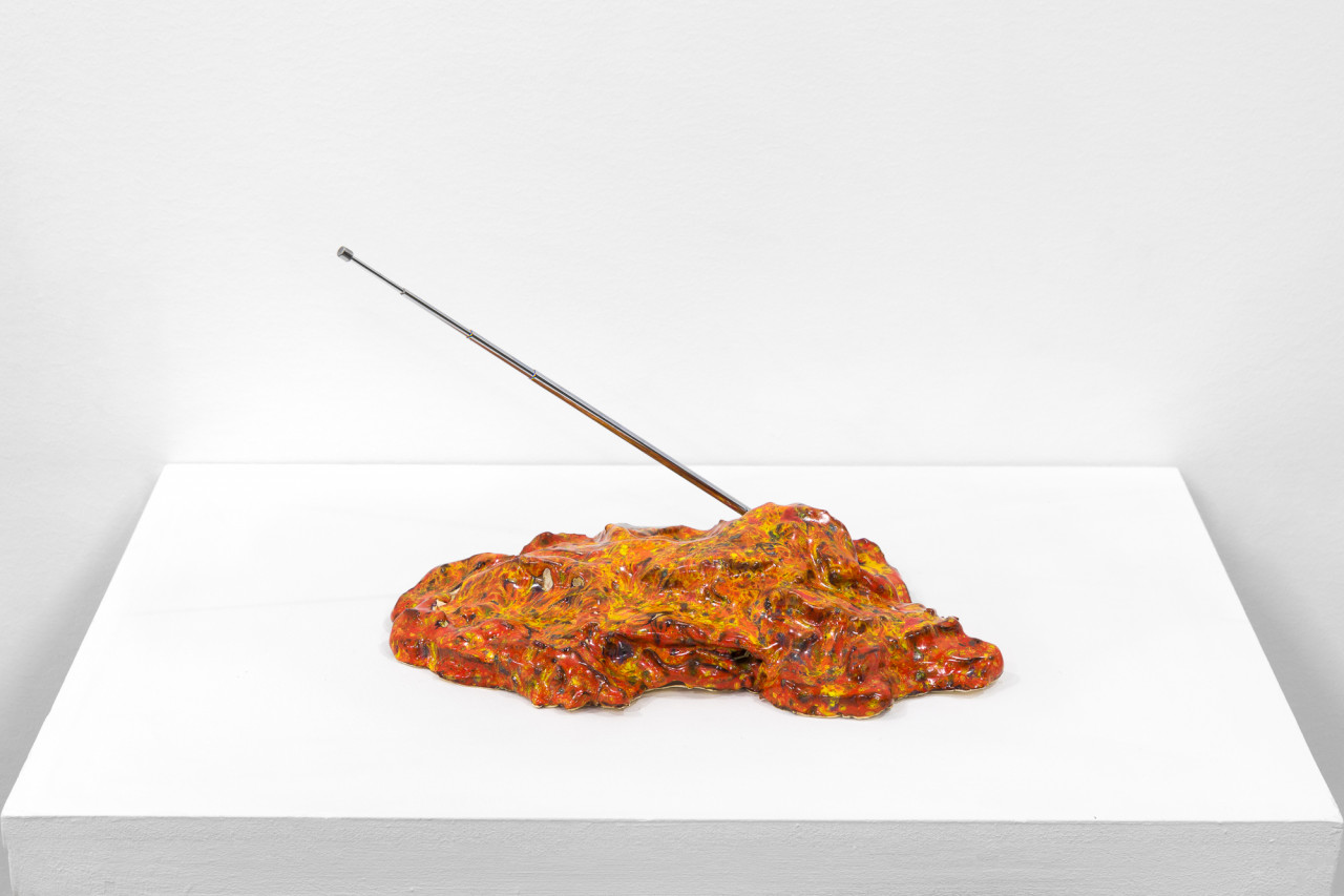 A multicolored object sits on a white base. A metal antenna extends out on a diagonal.