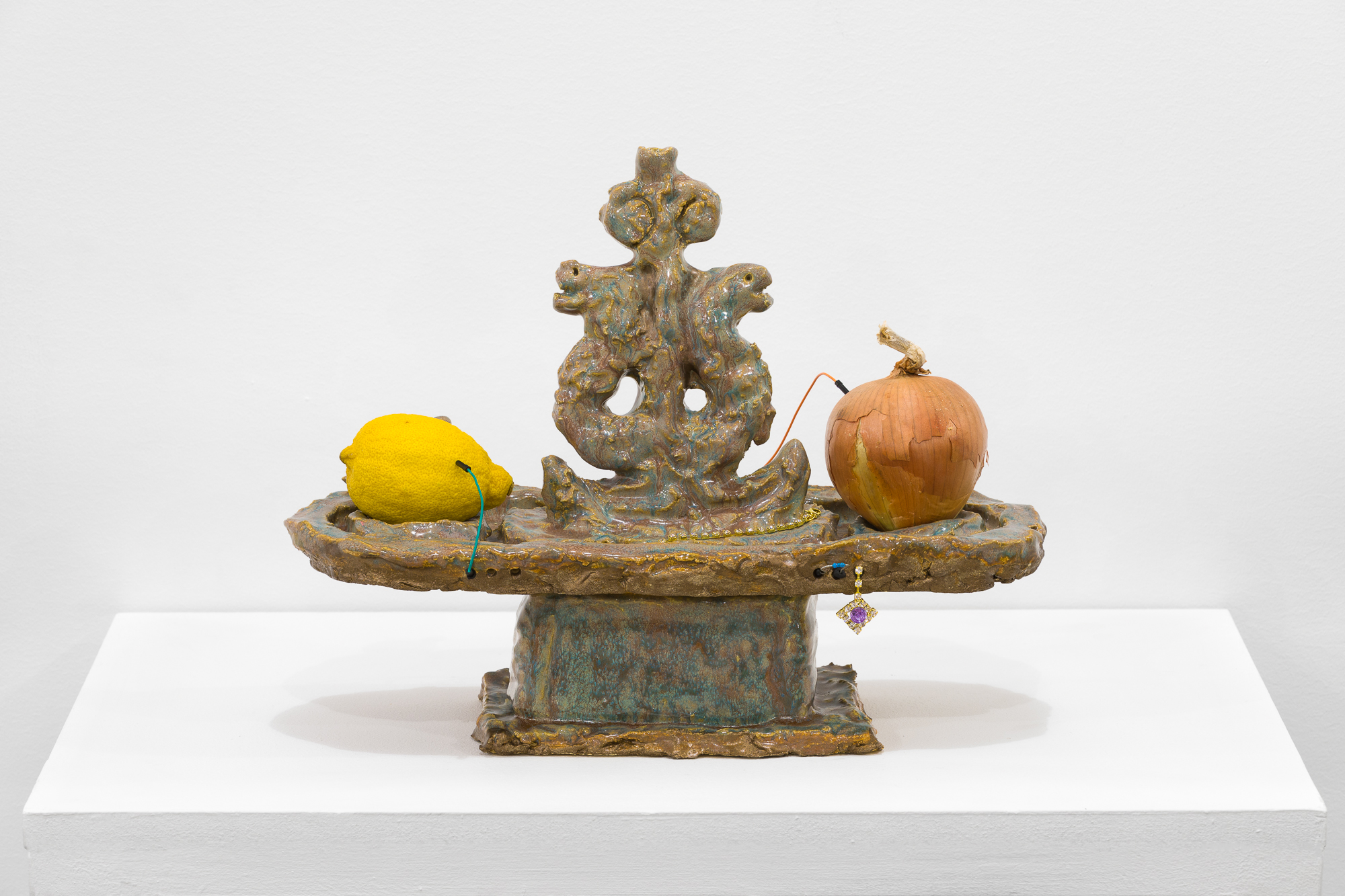A brown, blue, and tan object stands on a white base. On the flat surface of the object sit an onion and a lemon, each with wires connecting them to the object. Under the onion, a purple earring hangs down.