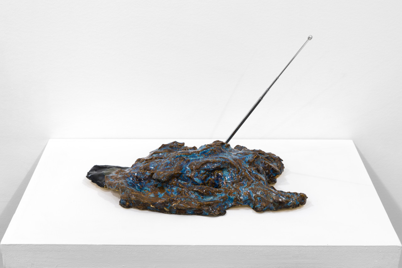 A blue, black and brown abstract object sits on a white base. A metal antenna sticks out of it on a diagonal.