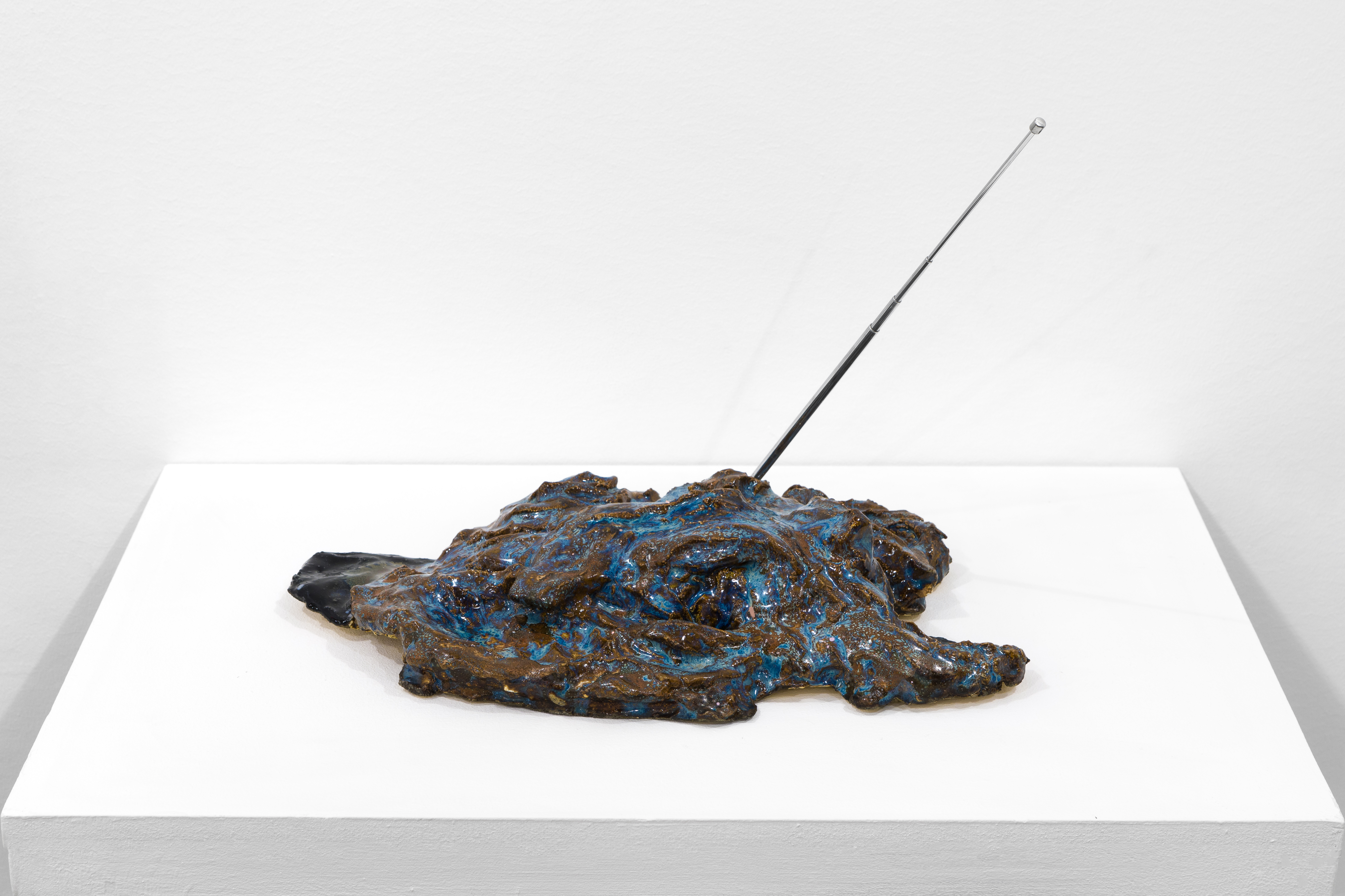 A blue, black and brown abstract object sits on a white base. A metal antenna sticks out of it on a diagonal.