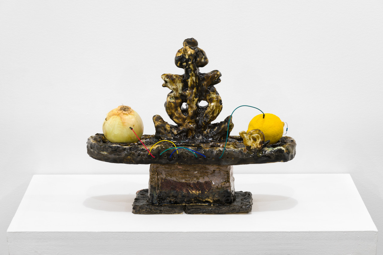 A brown object stands on a white base. On the flat surface of the object, an onion and a lemon are connected by a rainbow wire.