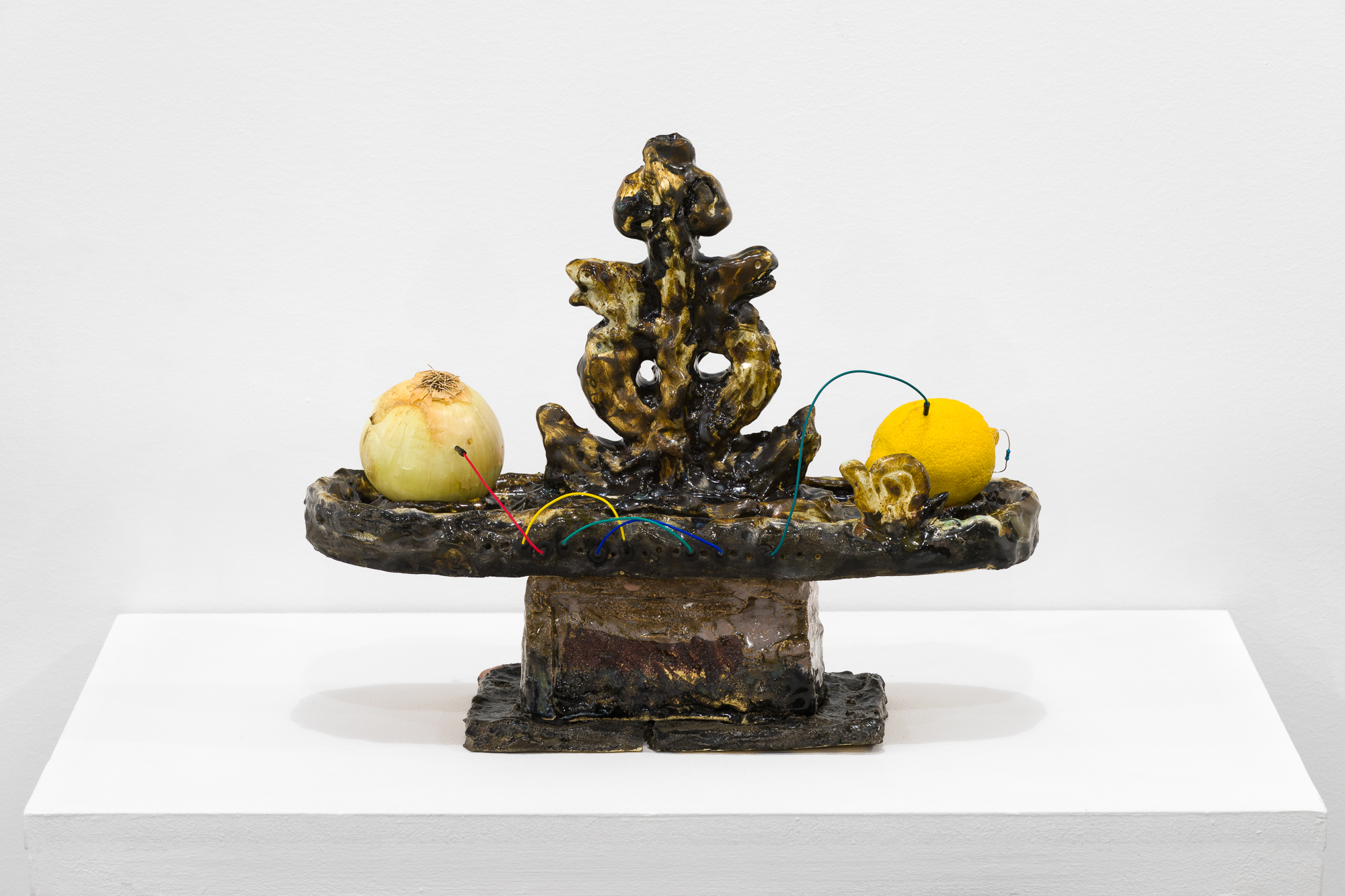 A brown object stands on a white base. On the flat surface of the object, an onion and a lemon are connected by a rainbow wire.