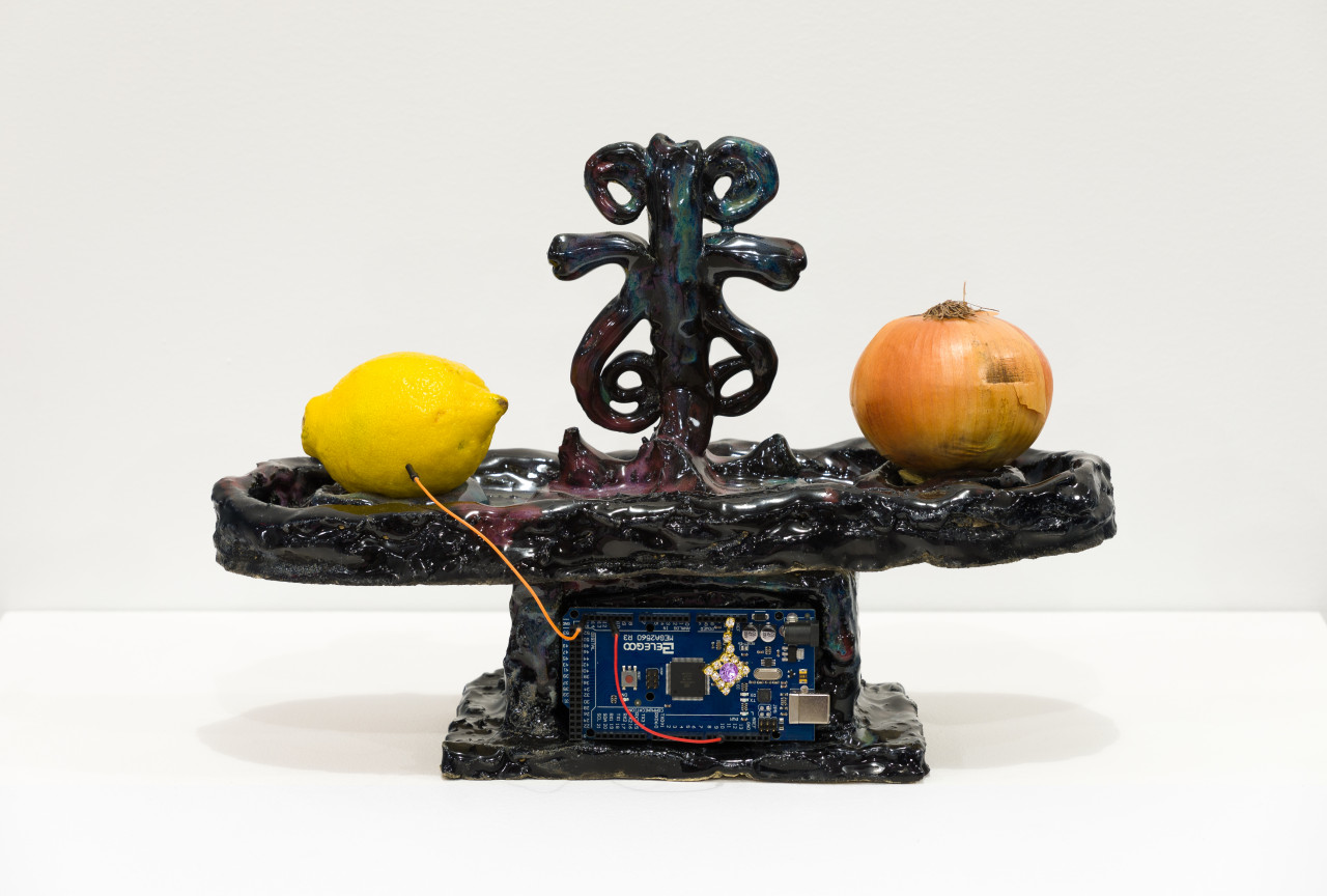 A glossy object with dark tones showing glimpses of bright blue and purple throughout is pictured on a white base. The object has a lemon and an onion balanced on either end, with electrical cords running from the produce to a chipboard at the object's center.