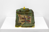 A yellow object painted with a green and grey cracked surface sits on a white base. There is a rectangular panel with wiring attached to the front. On top of the object sits a small yellow figure.