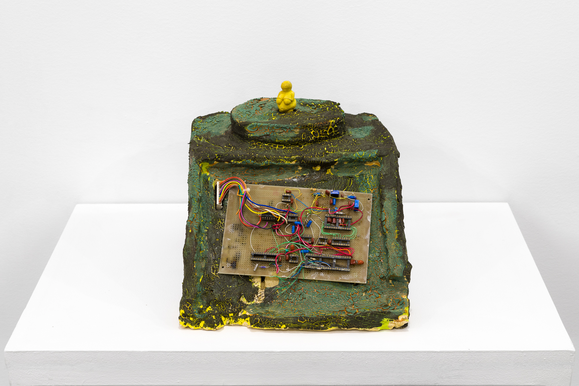 A yellow object painted with a green and grey cracked surface sits on a white base. There is a rectangular panel with wiring attached to the front. On top of the object sits a small yellow figure.