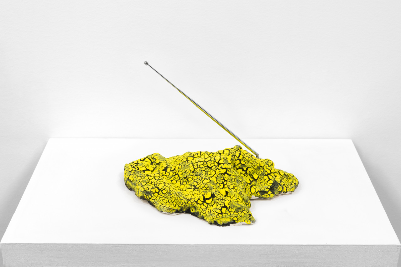 A yellow object with a cracked surface sits on a white base. A metal antenna extends out on a diagonal.