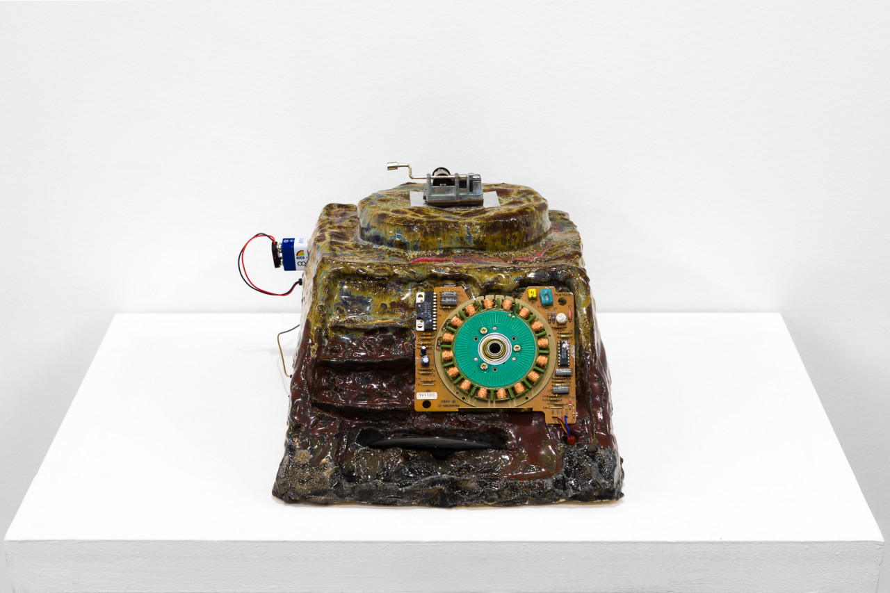 A brown and green object with a processing chip imbedded in its front sits on a white base. There is a battery with two wires sticking out the left side, and a small, silver crank device sitting on top.