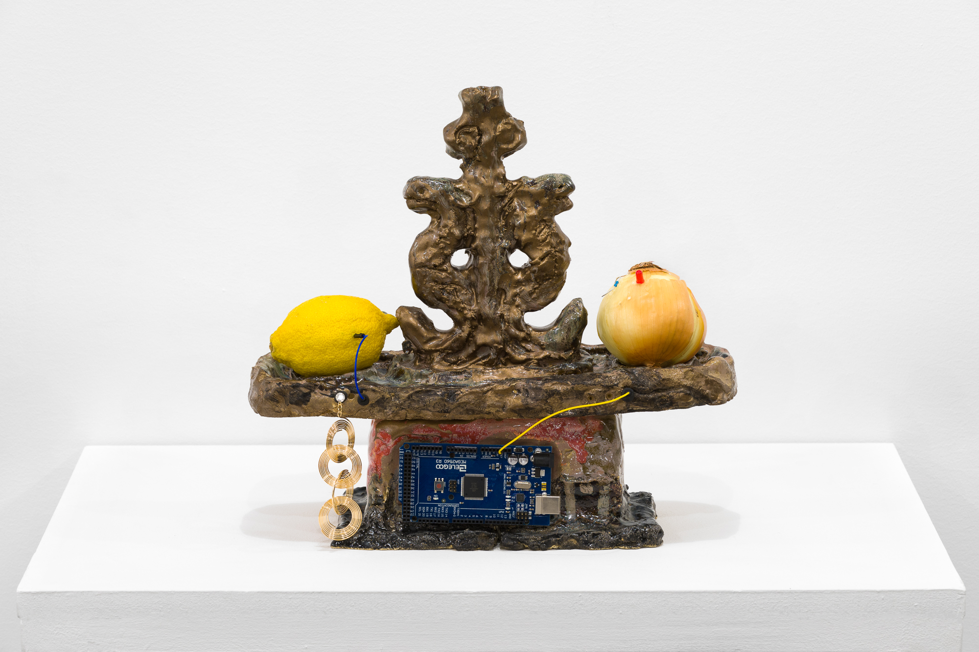 A brown and gold object stands on a white base. On the flat surface of the object sit an onion and a lemon, each connected to a wire. In the base of the object a processing chip is embedded. Under the lemon, a gold earring hangs down.