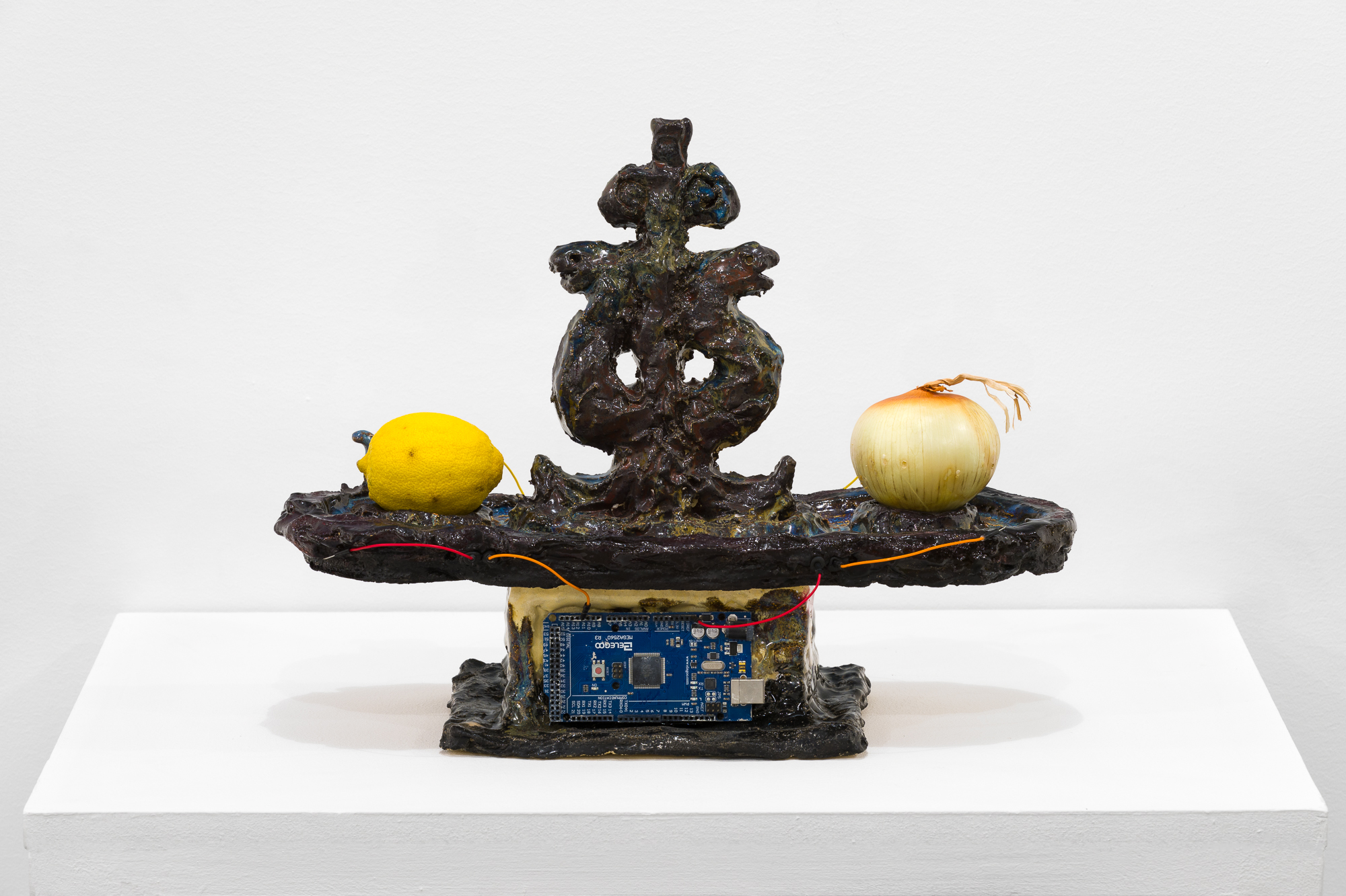 A brown object stands on a white base. On the flat surface of the object, an onion and a lemon are connected by a red and orange wire. In the base of the object, a processing chip is embedded.