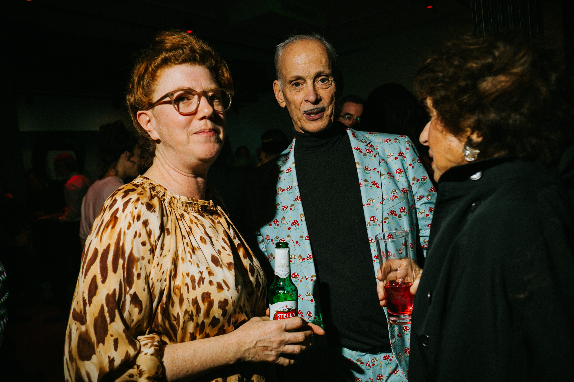 A color photo of three individuals standing together amidst conversation and festivities. The individual to the left and the person in the center both look towards the camera jovially.