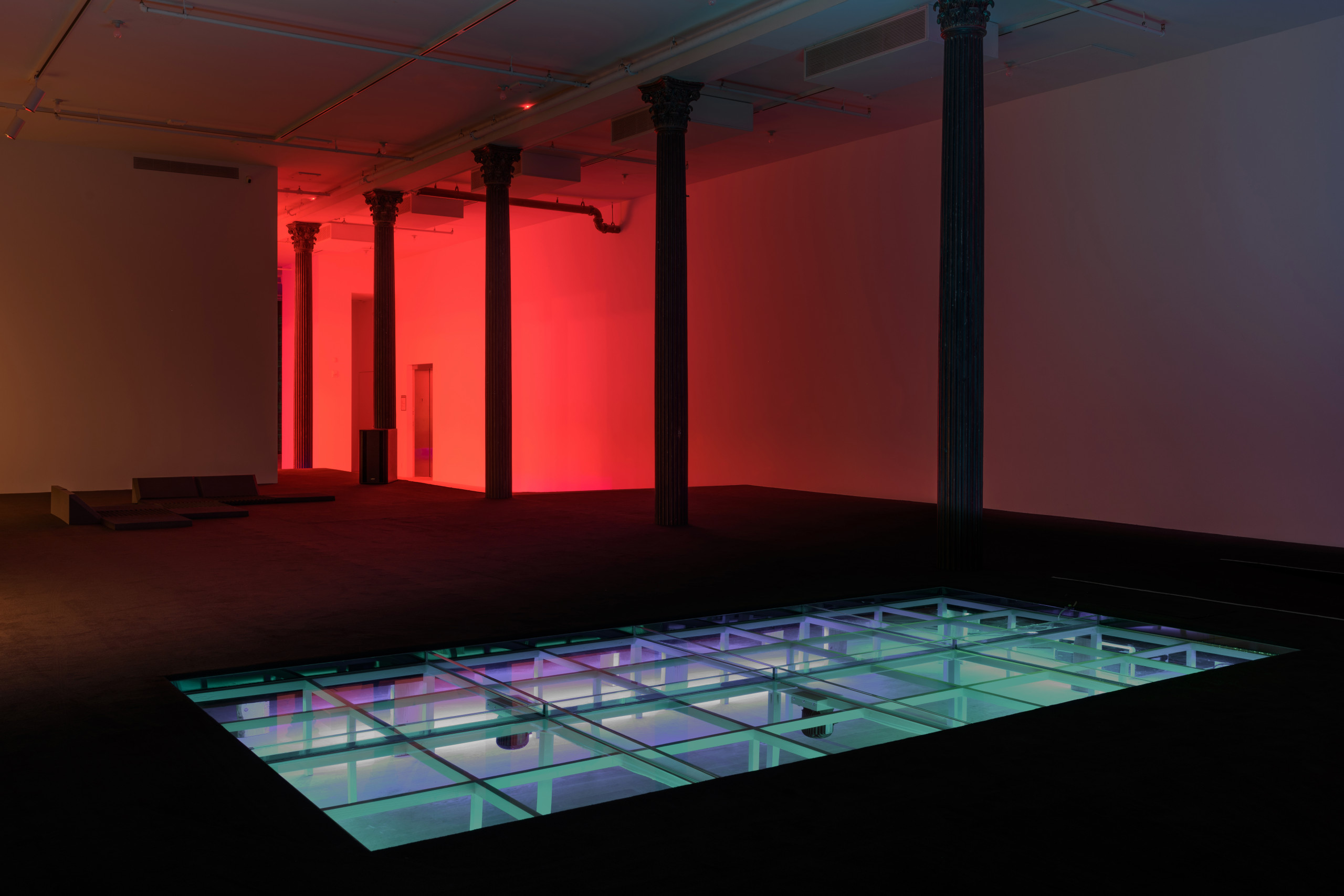 In a large dark room, a modular structure is recessed into a raised floor covered with black carpet. The structure is lit in bright blue light, and hosts open water. Red light shines in the back room.