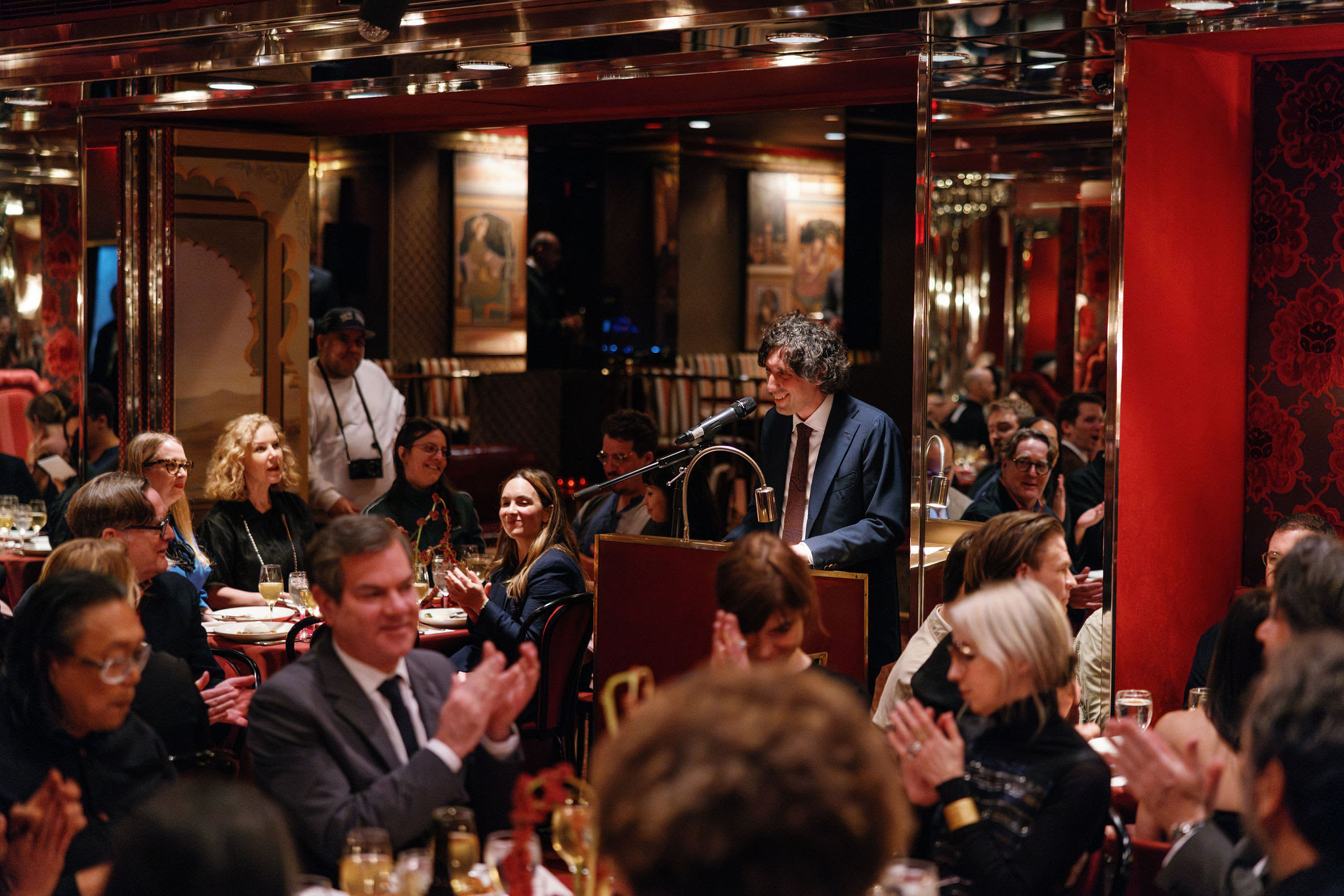 Jay Sanders stands at a podium in front of a microphone, smiling while mid-speech. Various gala attendees clap from their seats at circular tables arranged around the podium.