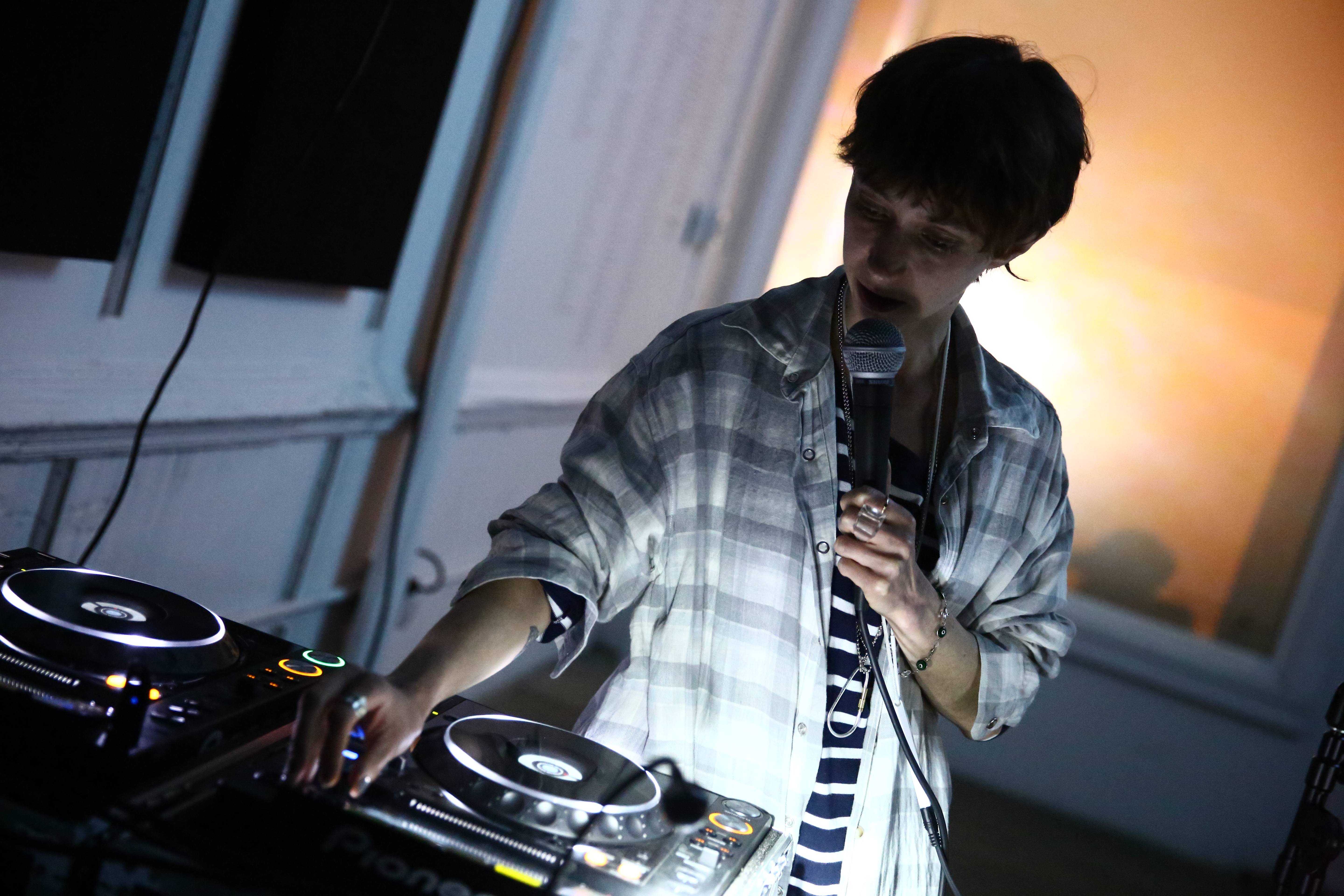 A person stands, holds a microphone to their mouth and adjusts a knob on a CDJ.