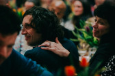 A color photo of two smiling individuals seated at a banquet table, the person to the right has her hand on another guest's shoulder. The background depicts several other guests out of focus.