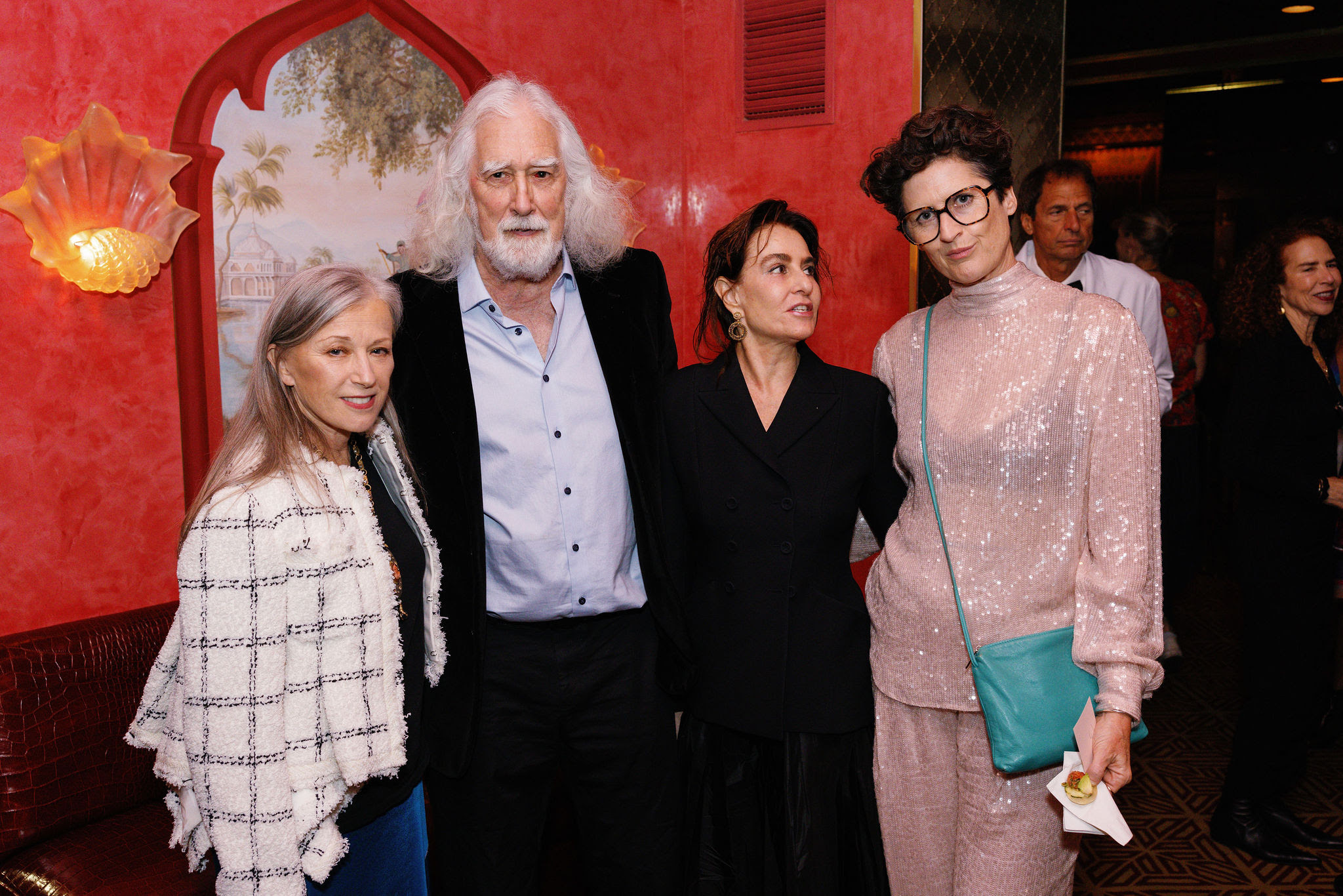 Cindy Sherman, Jack Hanley, Claudia Gould, and Alexandra Audler stand together and smile at the camera in front of a red wall with an illusionistic landscape view inside a painted pointed arch window. 