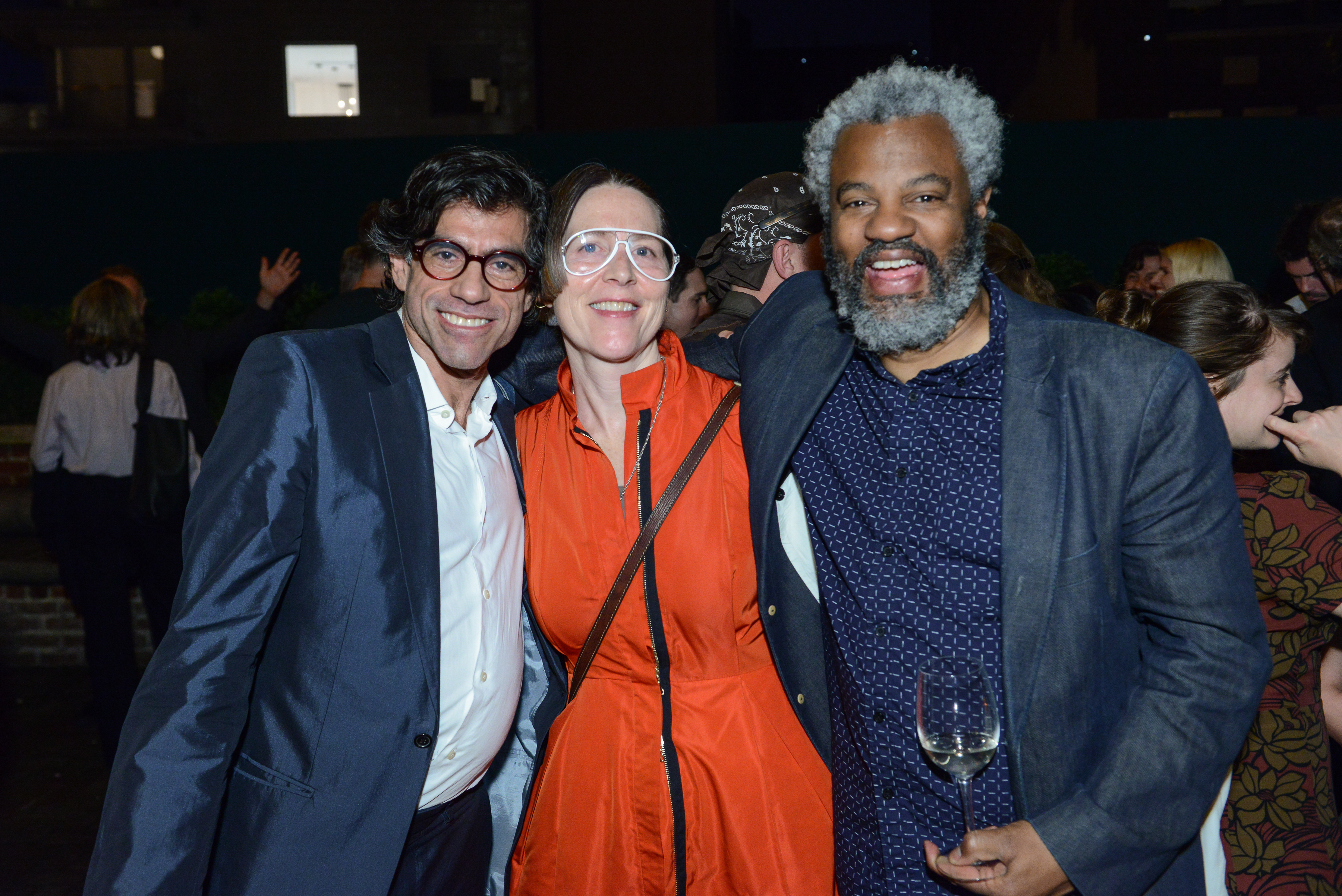 A color photo of three individuals smiling and posing together in an embrace for the camera. Other individuals engage with each other in the background of a dimly lit space.