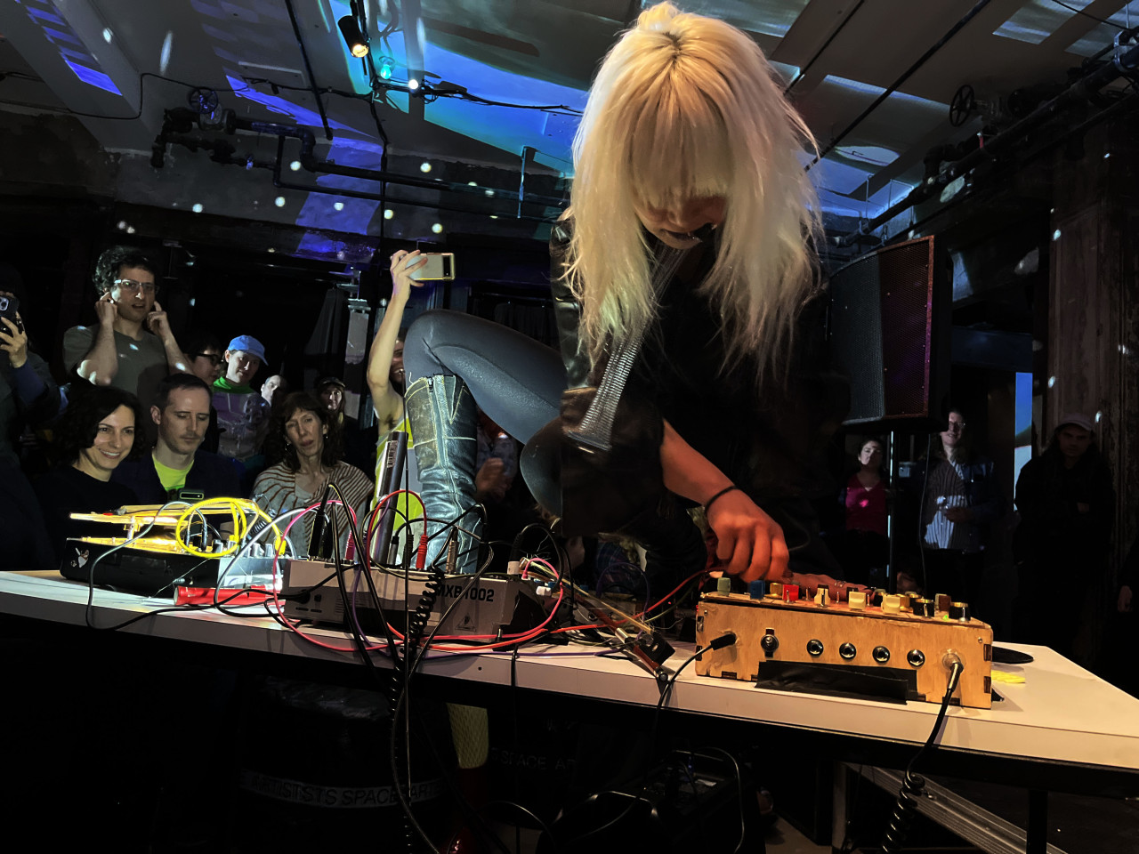 Photograph of a performer, leaning over a white table filled with audio equipment. The performer has their leg up on the table and use one hand to manipulate the equipment.