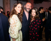 A color photo of three individuals standing together and posing for the camera before them. The background depicts other people engaging in festivities within a dimly lit space.