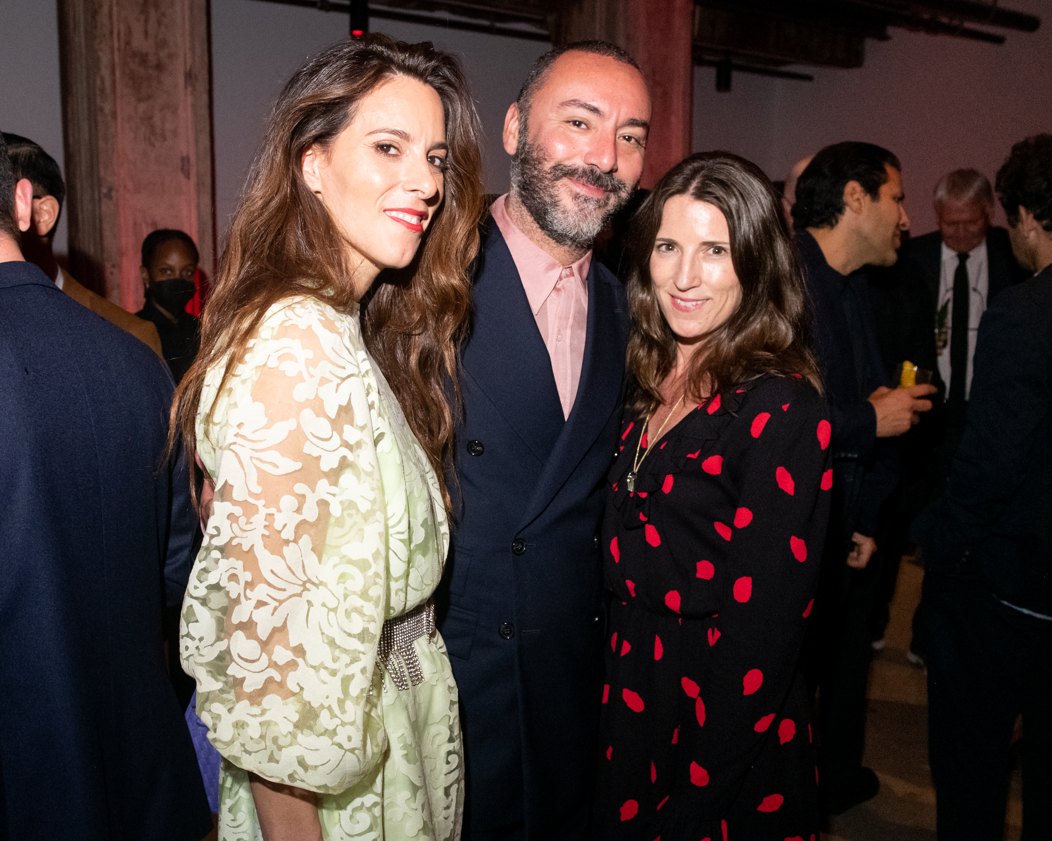 A color photo of three individuals standing together and posing for the camera before them. The background depicts other people engaging in festivities within a dimly lit space.