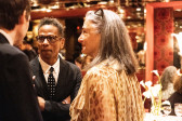 Color image of three or four individuals captured in the middle of a conversation. Two individuals have their back turned to the camera, while another person is facing the lens with arms crossed, actively listening. An elegant and decorative dinner venue can be seen in the background.