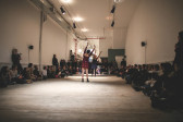 Two performers stand in the middle of a room, facing each other, with their arms raised, leaning slightly to one side. The audience sits on the perimeter of the room, forming a circle around them.