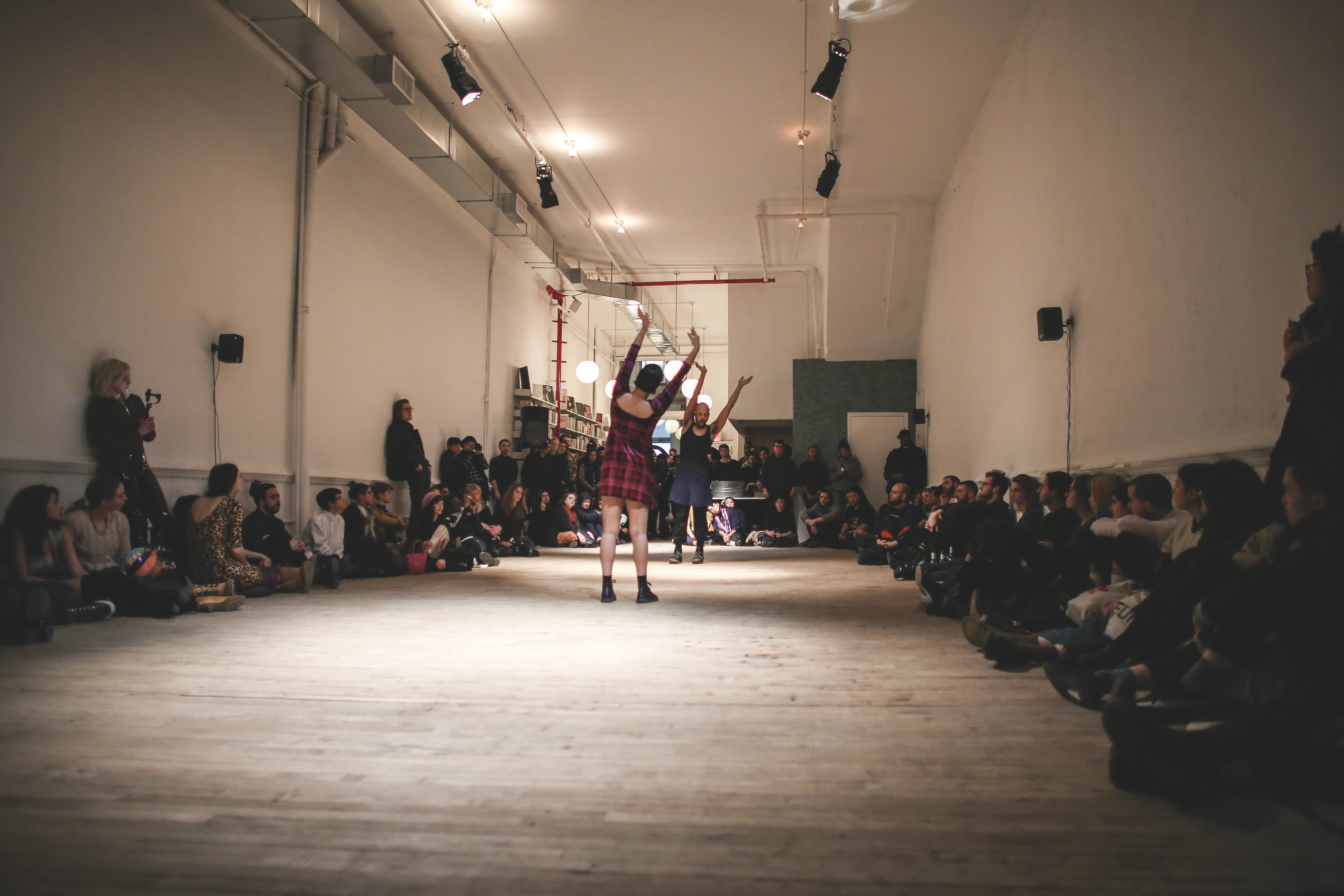 Two performers stand in the middle of a room, facing each other, with their arms raised, leaning slightly to one side. The audience sits on the perimeter of the room, forming a circle around them.