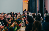 A color photo of several individuals seated at banquet-style dining tables. Individuals are clapping and looking around the large reception space.