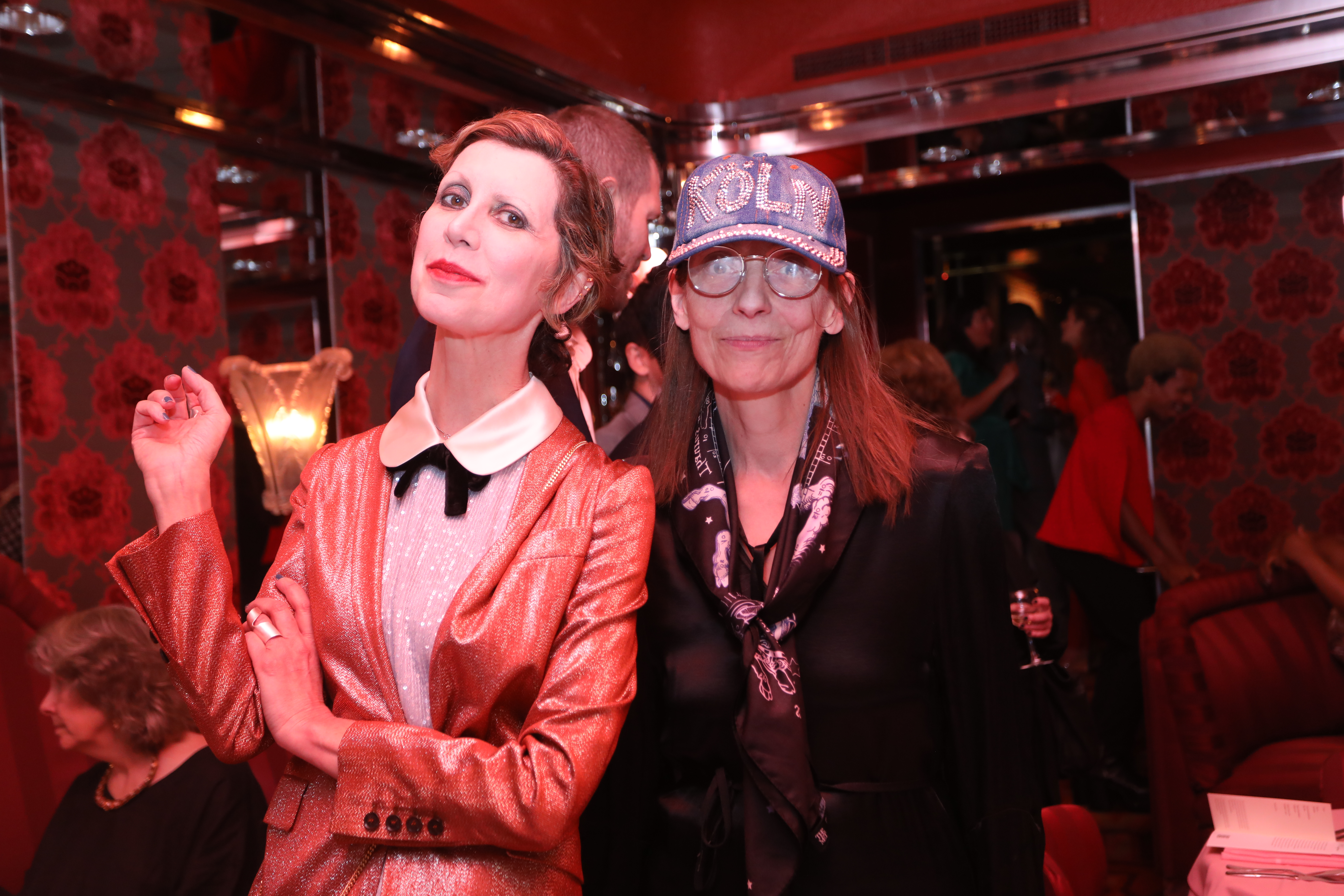 Color image of two individuals smiling jovially towards the camera. A dimly lit interior space with red flocking on the walls can be seen in the background.