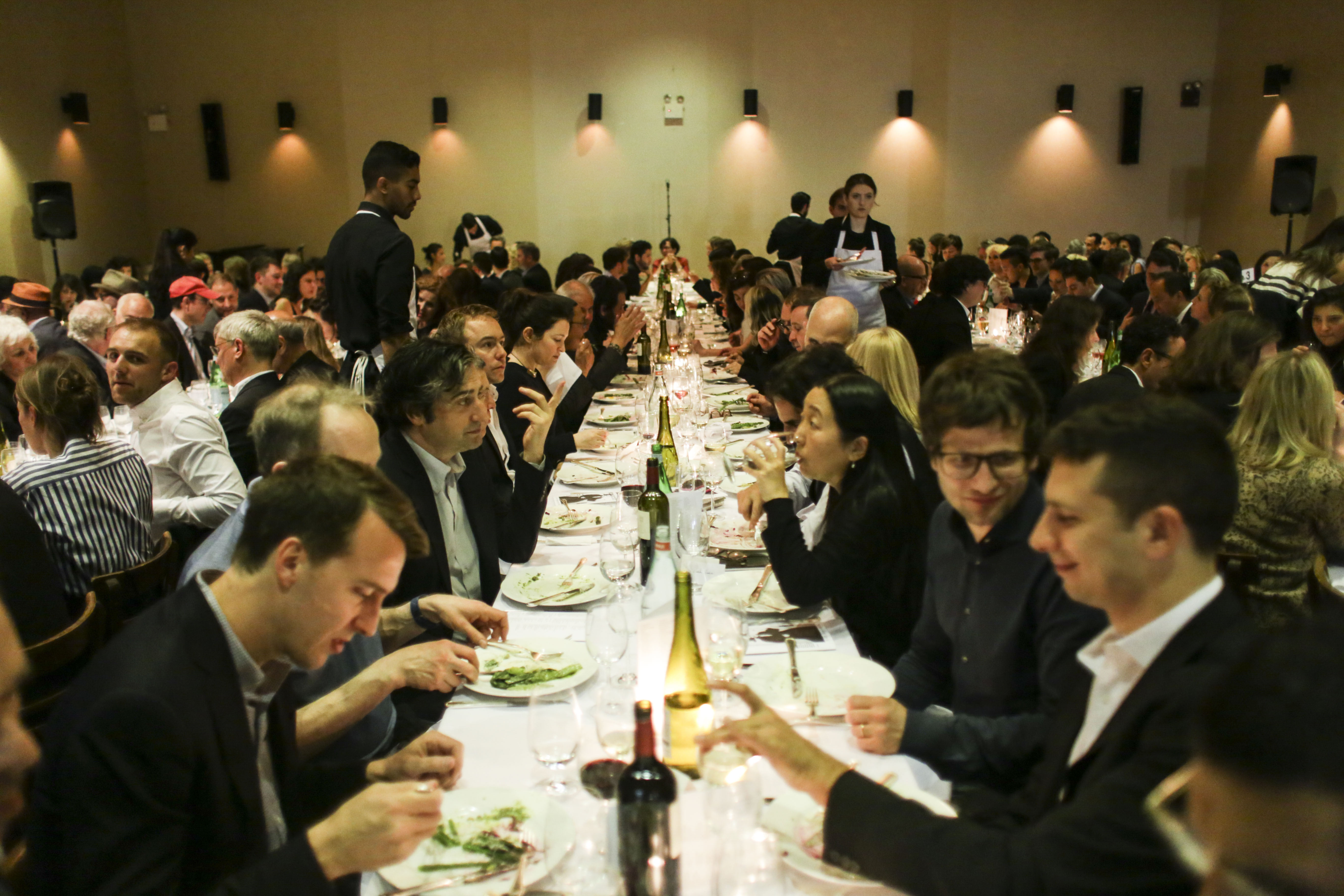 A large room with numerous people seated at long, rectangular tables. Many are eating, talking, and drinking wine. Several waiters walk between the tables.