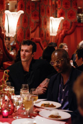 Two men, Drake Carr and George Wells, sit around a circular dining table, both gazing out while listening to a speaker who is out of frame. Carr rests his elbow on the back of his chair.