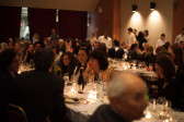 A photo of a room full of people seated at three long, rectangular tables. Each table has numerous glasses of wine, sparkling water, candles, and empty plates of food. In the foreground, people are talking to each other and smiling.