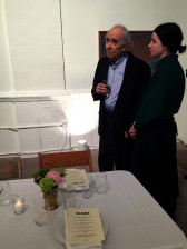 Two people stand at the foot of an empty table, mid-conversation, looking off-camera to the left. The individual on the left holds a glass of wine.
