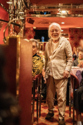 Color image of an individual standing and smiling jovially towards the camera. A podium can be seen to the left of the image, and a variety of seated party guests are also imaged within the elegant and decorative venue.