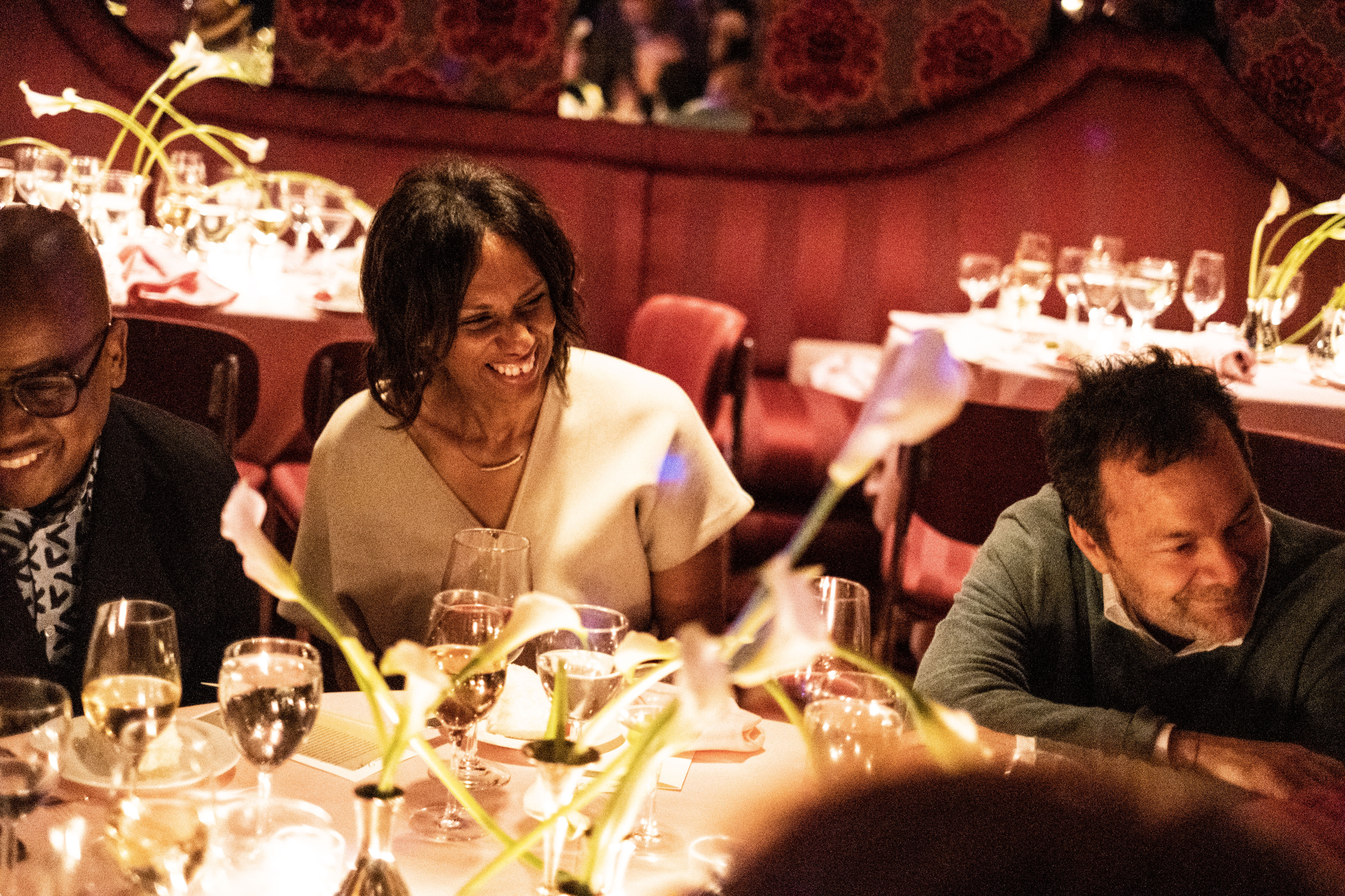 Color image of three individuals captured as they engage with other guests at their table. A dimly lit and elegant dinner venue can be seen in the background.