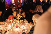 Color image of four individuals crowded together as they look at something on a cellphone. Two individuals in the group are seated at a dinner table, where other guests are also engaged in conversation. A dimly lit and elegant venue full of other individuals can be seen in the background.
