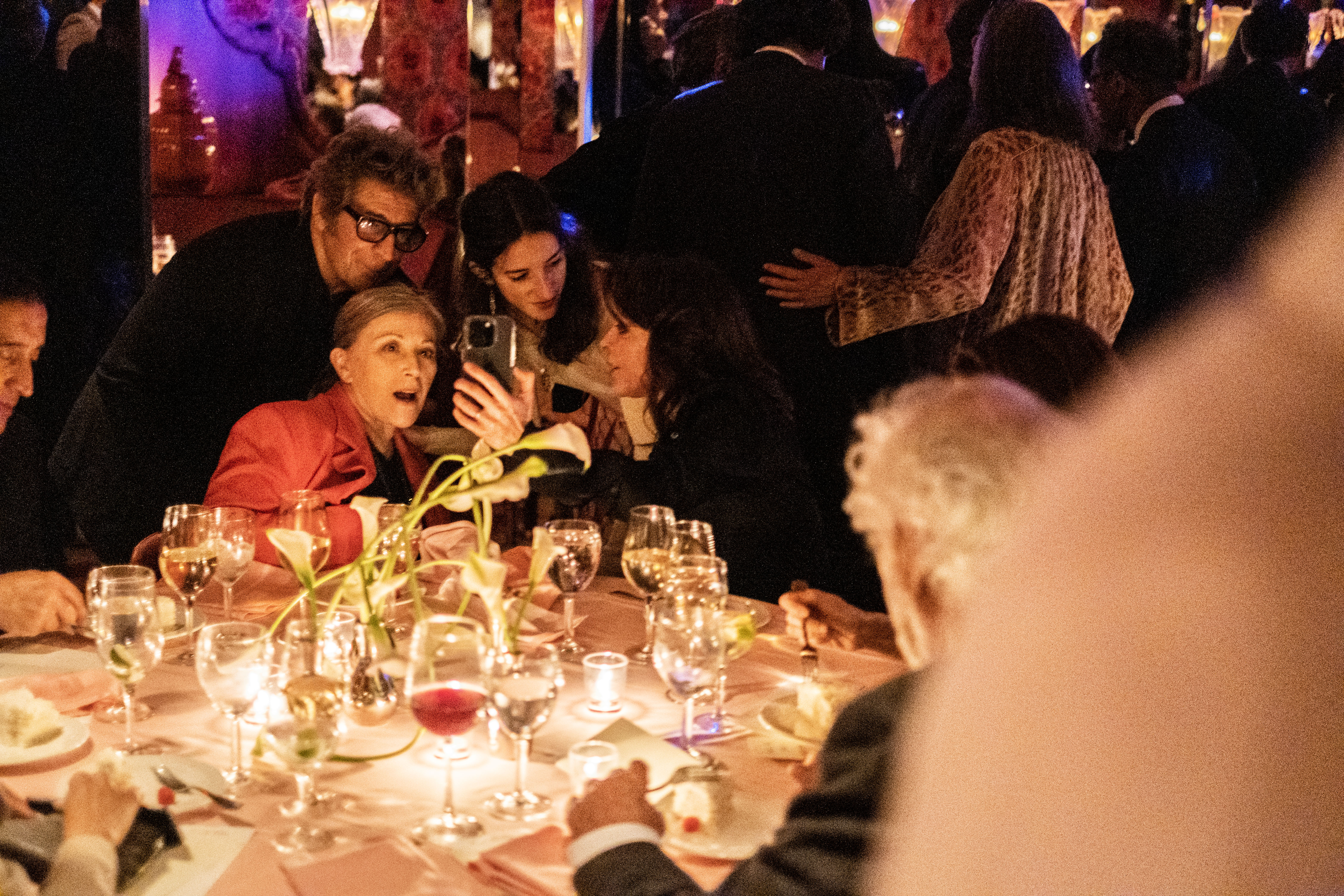 Color image of four individuals crowded together as they look at something on a cellphone. Two individuals in the group are seated at a dinner table, where other guests are also engaged in conversation. A dimly lit and elegant venue full of other individuals can be seen in the background.