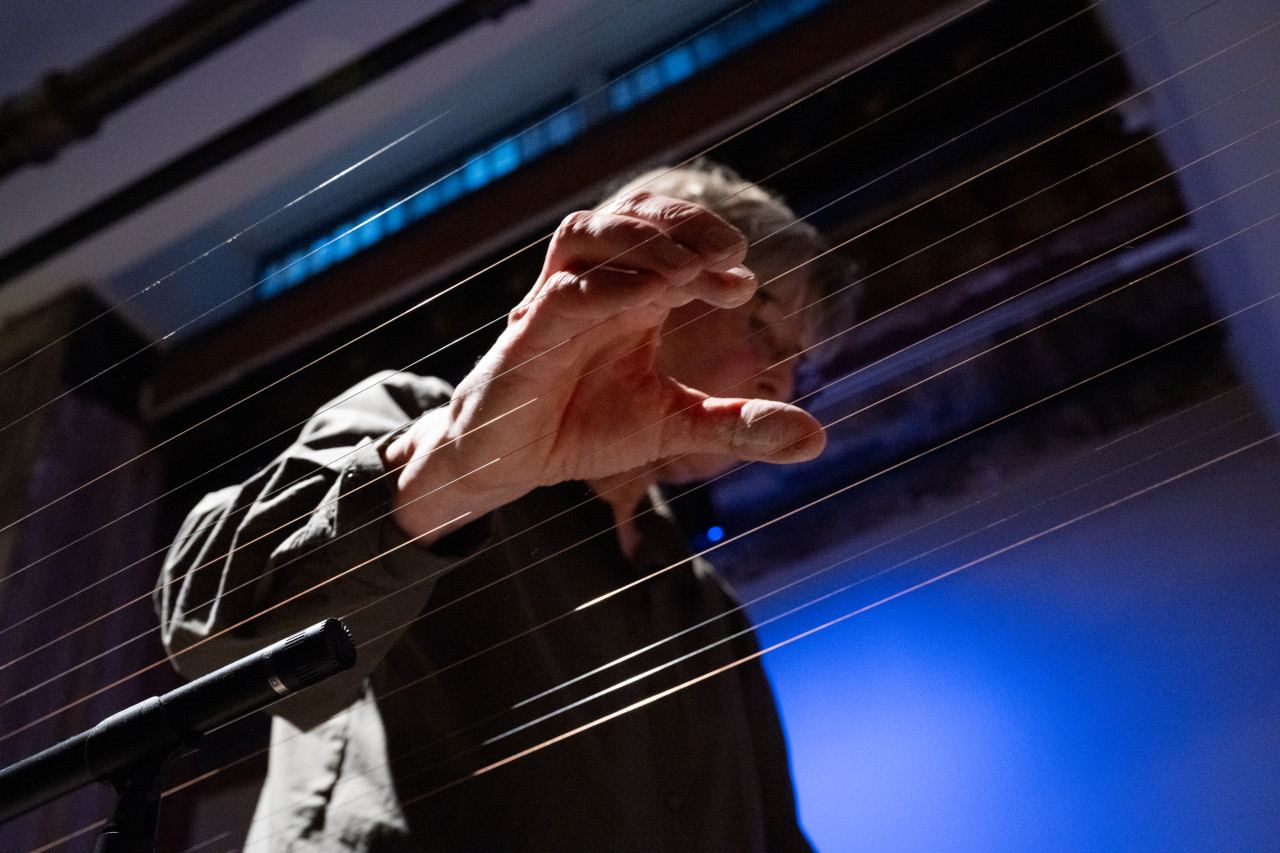 A photograph taken from a low angle, looking up at a performer whose hand is poised on top of a long set of instrumental wires that run across the image.