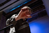A photograph taken from a low angle, looking up at a performer whose hand is poised on top of a long set of instrumental wires that run across the image.
