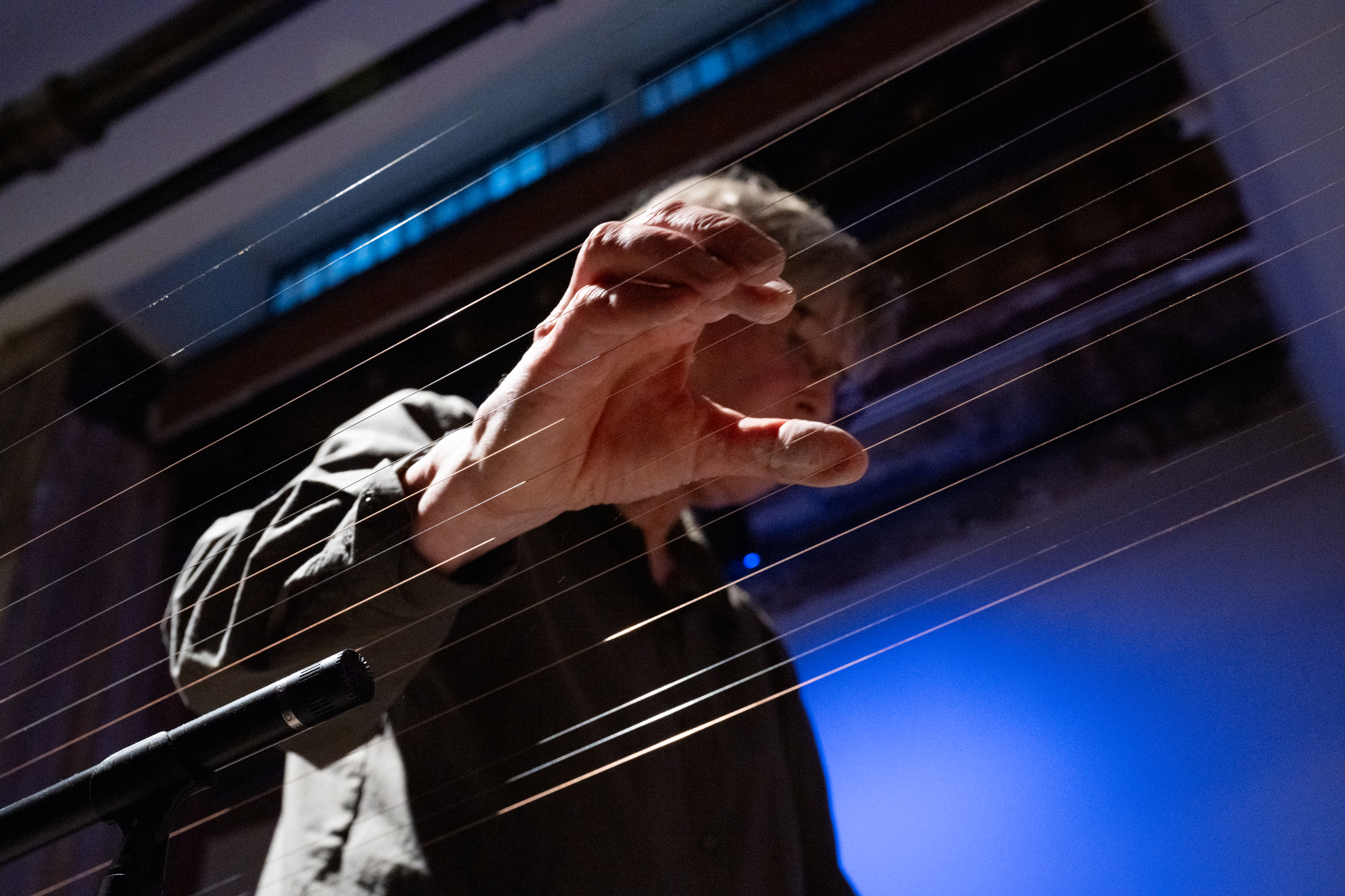 A photograph taken from a low angle, looking up at a performer whose hand is poised on top of a long set of instrumental wires that run across the image.